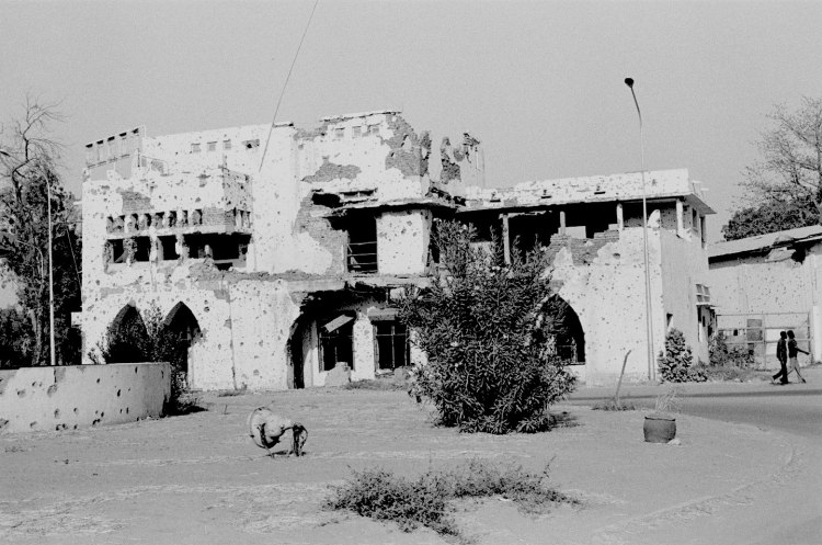 building with bullet holes two men walking to the right and a large shrub in the centre