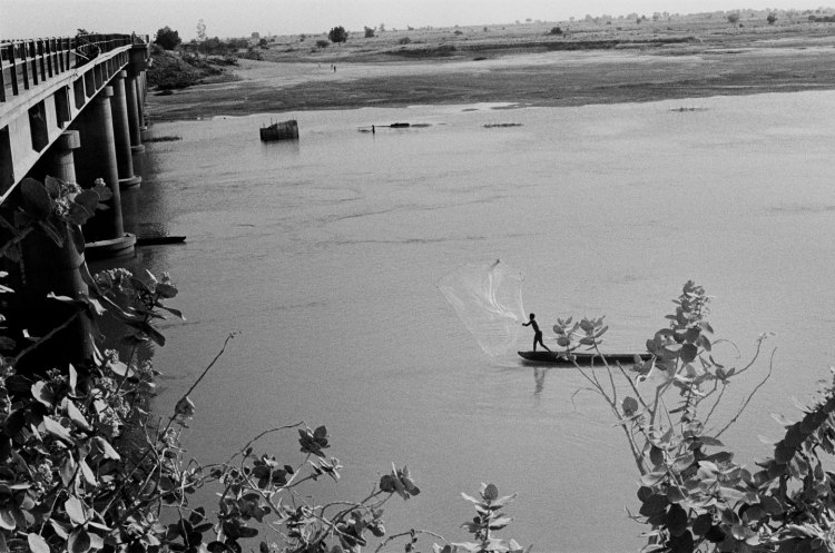 fisherman casting his net on fishing boat river chari with bridge to left and chad landscape in background africa 1982
