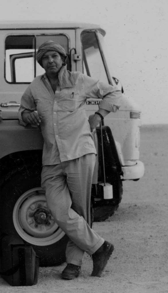 john howe with scarf round head standing with a truck in the desert north africa