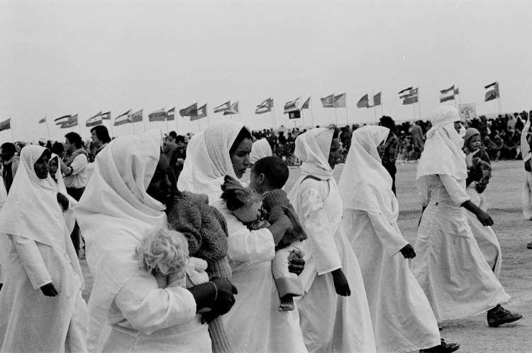 women marching in white robes and headdresses with babies crowd and flags behind polisario ten year celebration western sahara 1986