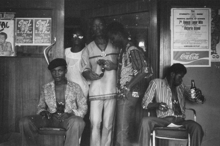 group of guys wearing cool 70s clothing and shades and drinking beer standing at double doors in lagos nigeria 1978 with posters