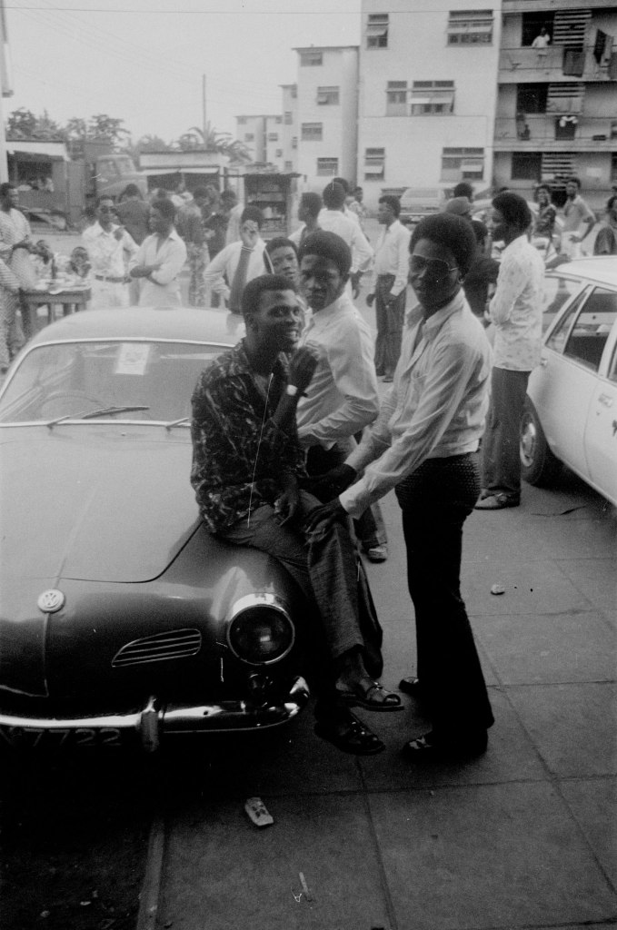 boys wearing cool clothes hanging out by an old volkswagen in lagos nigeria with a crowd in the background and apartment blocks behind 1978