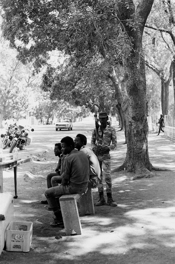 three men seated on bench and soldier with gun standing motorciycles and car in background dappled sunlight ndjamena chad 1982