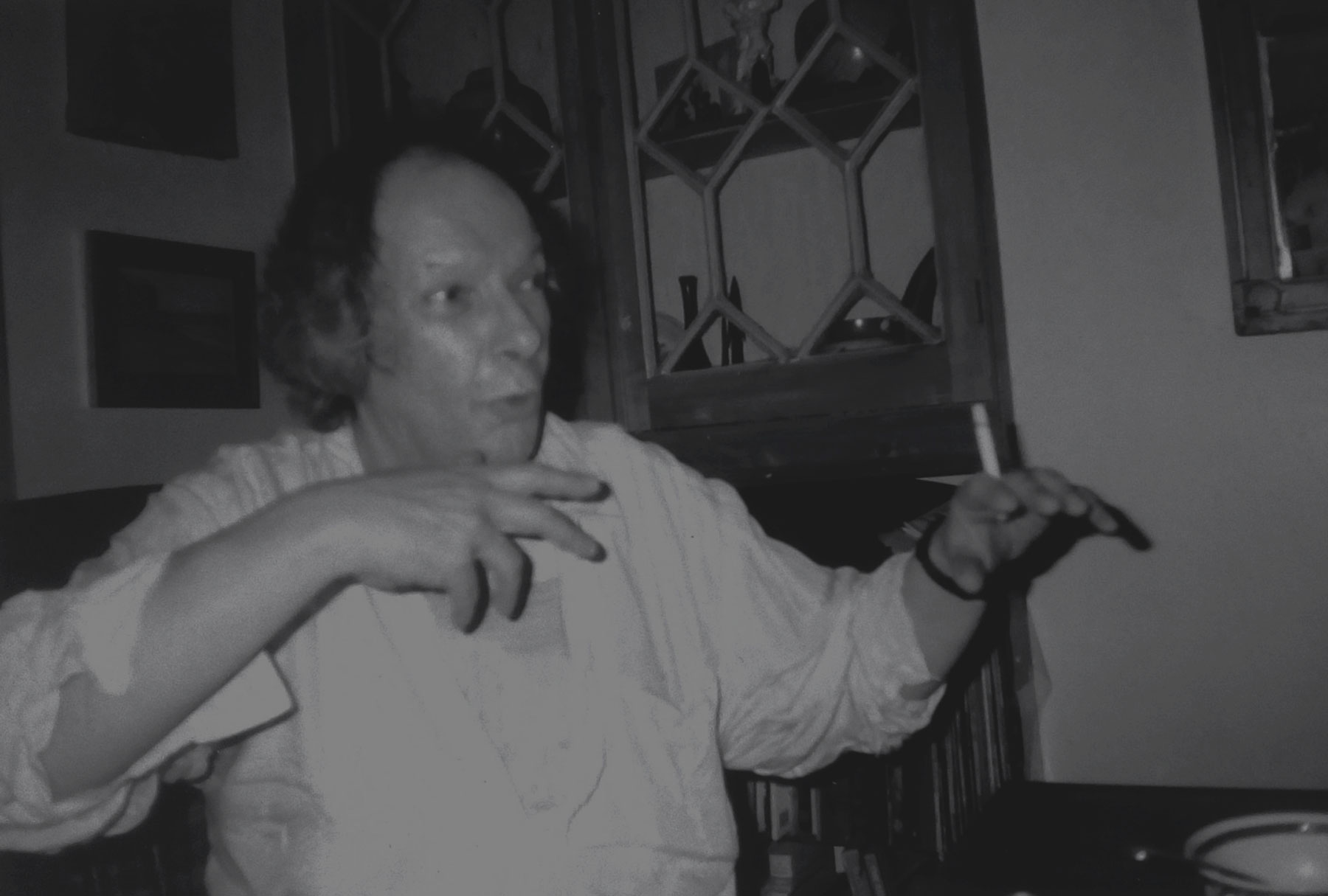 john howe describing and making hand gesture while smoking in sitting room with pictures behind