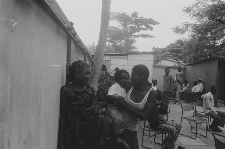 women standing in the yard under a palm tree with a baby and men sitting around in the background nigeria 1978
